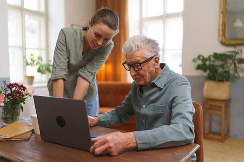 Enkelin bringt Großvater bei, wie man einen Computer, ein Notebook benutzt. Junge Frau, Betreuerin zeigt Seniorin etwas auf einem Laptop. - HPIF35081