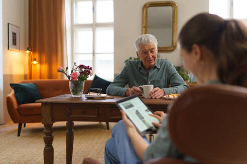 Eine Pflegekraft liest eine Online-Zeitung auf einem digitalen Tablet, während sie sich um einen älteren Mann in seinem Haus kümmert. - HPIF35085