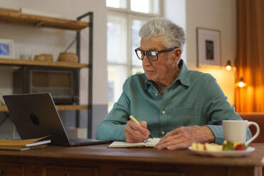 Älterer Mann schreibt Notizen in sein Tagebuch, mit geöffnetem Computer. Älterer Mann isst Snack vor einem Computer. - HPIF35090