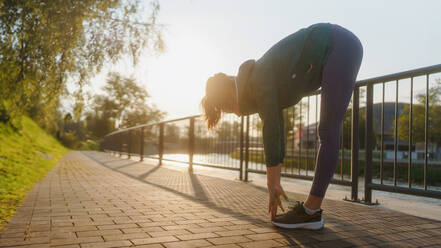Junge Läuferin dehnt Arme und Beine vor ihrem Morgenlauf in der Stadt. Fitness-Mädchen in Sportkleidung bereitet sich auf das Abendtraining vor. Outdoor-Workout-Konzept. Sillhouette mit Morgenlicht. - HPIF35108
