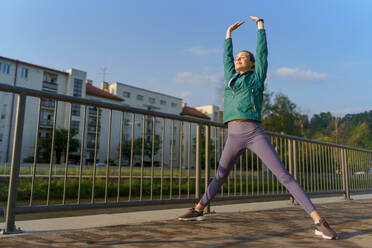 Young female runner stretching before her early morning run in the city. Fitness girl in sportswear preparing for evening exercise. Outdoor workout concept. - HPIF35118