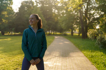 Porträt einer jungen, hübschen Läuferin in blauer Jacke, deren Haare im Wind wehen, die durch den Stadtpark joggt. Abendliches Training in der Stadt. Outdoor-Workout-Konzept. - HPIF35135