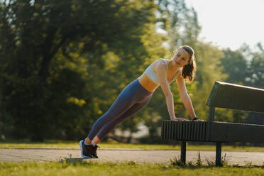 Full lenght portrait of beautiful fitness woman doing push-ups against park bench. Workout at the park. Park bench excercises. - HPIF35146