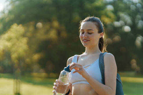 Mittlere Einstellung eines sportlichen Mädchens in Sportkleidung mit Yogamatte auf der Schulter, das aus einer wiederverwendbaren Wasserflasche trinkt. - HPIF35148