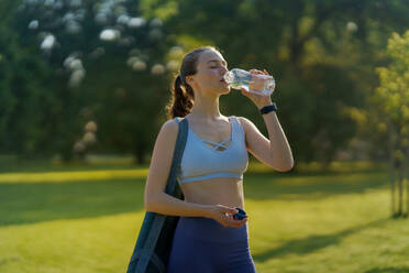 Side view of beautiful athletic girl in sportswear with yoga mat on shoulder drinking water from bottle heading for a pilates workout. - HPIF35149