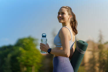 Seitenansicht eines schönen, sportlichen Mädchens in Sportkleidung mit Yogamatte auf der Schulter und Wasserflasche auf dem Weg zu einem Pilates-Workout. - HPIF35151