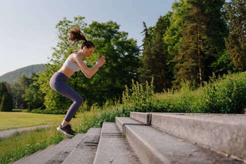 Sportliche Frau macht hüpfende Kniebeugen auf einer Treppe im Stadtpark. Weibliche Athletin in Sportkleidung beim Fitnesstraining in der Stadt. Gesundes Lifestyle-Konzept. - HPIF35153