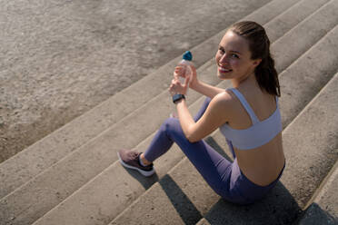Draufsicht auf junge Fitness-Frau in Sportkleidung, die sich nach dem Training in der Stadt ausruht. Schöne sportliche Frau, die nach dem morgendlichen Training auf einer Betontreppe Wasser trinkt. - HPIF35164