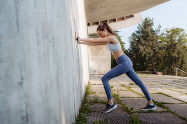 Seitenansicht einer jungen sportlichen Frau in Sportkleidung, die im Freien trainiert. Fitness-Frau, die sich gegen eine Betonwand in der Stadt streckt. Konzept für einen gesunden Lebensstil. Street Workout. - HPIF35167