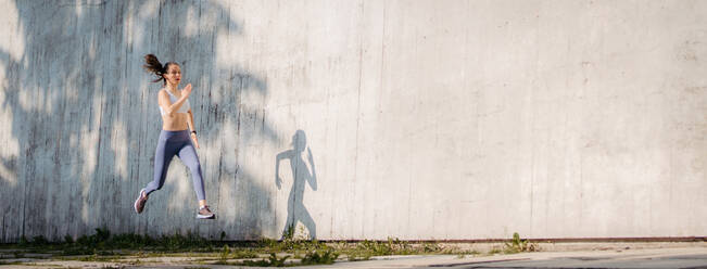 Sportlerin in Sportkleidung beim Training in der Stadt. Sie läuft vor einer Betonwand, die Schatten wirft. Banner mit Kopierbereich. - HPIF35180