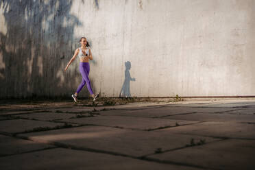 Sportlerin in Sportkleidung beim Training in der Stadt. Sie läuft vor einer Betonwand, die Schatten wirft. Banner mit Kopierbereich. - HPIF35181