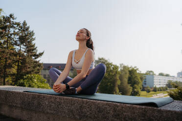 Front view of young sporty woman doing breathing exercises on gym mat after workout in the city. Woman meditate outdoors, in the park. - HPIF35194