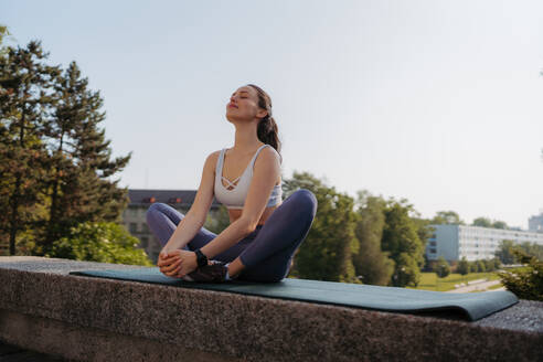 Frontansicht einer jungen sportlichen Frau, die nach dem Training in der Stadt Atemübungen auf einer Gymnastikmatte macht. Frau meditiert im Freien, im Park. - HPIF35194