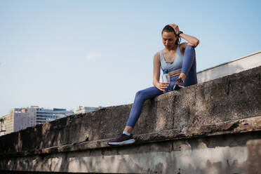 Junge Fitness-Frau in Sportkleidung, die sich nach einer harten Trainingseinheit in der Stadt ausruht. Sportliche Frau, die nach dem morgendlichen Training Luft holt und Wasser trinkt. - HPIF35201