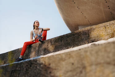 Young fitness woman in sportswear resting after hard workout session in the city. Sporty woman catching breath and drinking water after morning excercise. - HPIF35203