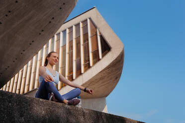 Low angle shot of young sporty woman doing breathing exercises after workout in the city. Woman meditate outdoors, sitting on concrete stairs in the park. - HPIF35207