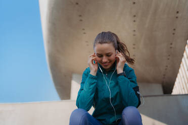 Young fitness woman listening to music with headphones, resting after workout session in the city. Beautiful sporty woman enjoying sunrise after morning excercise. - HPIF35209