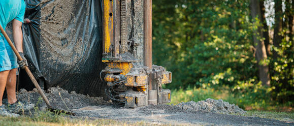 Close up of well drilling rig boring dowin into the earth. A rotary drill rig using bits to bore into ground and loosing the soil and rocks. Worker shoveling rubble. Drilling machine, equipment using water to cool down bits. Mud and muddy water is spitting out when drilling borewell. Banner with copy space. - HPIF35227