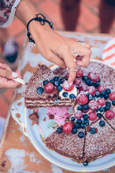 Nahaufnahme einer weiblichen Hand, die ein Stück nackten Schokoladenkuchen mit Sahne und Beeren auf einer Sommerparty im Freien hält - ADSF50280