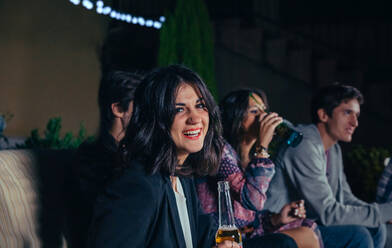 Porträt einer lächelnden jungen Frau, die ein Bier in der Hand hält, während sich ihre Freunde auf einer Party im Freien unterhalten. - ADSF50281