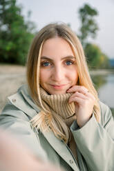 Positive junge Frau in Mantel und Schal, die in die Kamera schaut, während sie vor einem unscharfen Hintergrund eines Parks steht - ADSF50287