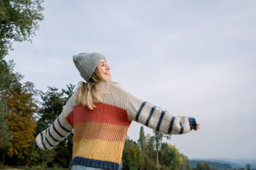 Von unten der jungen Frau in warmen Pullover und Strickmütze stehend gegen grüne Bäume und Wolke beim Genießen sonnigen Tag auf dem Lande auf Winterzeit - ADSF50289