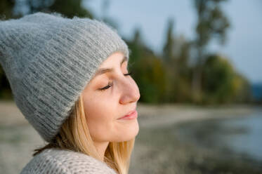 Side view of young woman in warm cozy sweater and hat, breathes clean air, looks at the sky with a smile and closed eyes, walks with pleasure in the autumn forest. people health content - ADSF50291