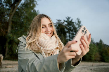 People and wanderlust. Overjoyed satisfied young woman makes selfie portrait, spends free time on beautiful lake, wears raincoat, stands on coast alone. Recreation and trip. Background trees - ADSF50292