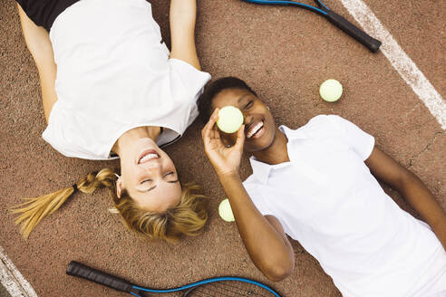 Portrait of two playful young beautiful women with tennis clothes and rackets laying on a tennis court. Two friends sharing a morning of sportive activity playing a tennis match. - ADSF50305