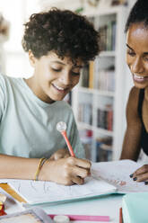 Cheerful woman helping daughter doing homework. Happy mother assisting her daughter with school work in living room. - ADSF50309