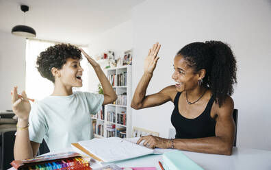 Fröhliche Frau, die ihrer Tochter bei den Hausaufgaben hilft. Mutter und Tochter schütteln sich die Hände, während sie im Wohnzimmer Hausaufgaben machen. - ADSF50310
