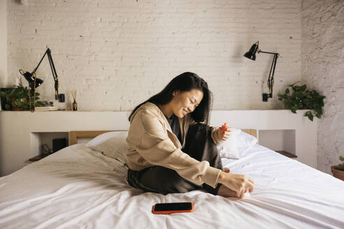 Junge chinesische fröhliche Frau macht Pediküre auf dem Bett zu Hause - ADSF50325
