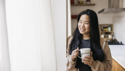 Junge schöne Frau mit einem Becher Kaffee trinkend neben einem Fenster in einer Küche. Lächelnde Frau, die eine Kaffeepause zu Hause genießt. - ADSF50327