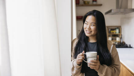 Young beautiful woman with a mug drinking coffee next to a window in a kitchen. Smiling woman enjoying a coffee break at home. - ADSF50327