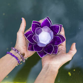 Blick von oben auf die Hände, die einen lila Glaslotus über Wasser halten. Yoga und Meditation Konzept. - ADSF50331
