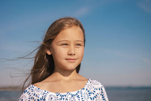 Nahaufnahme eines jungen Mädchens mit vom Wind zerzaustem Haar, das ein blau gemustertes schulterfreies Oberteil trägt und vor einem klaren blauen Himmel in die Ferne blickt - ADSF50343