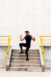 Full body side view of cheerful sportsman listening music over headphones and demonstrating muscles while standing on concrete staircase in city - ADSF50345