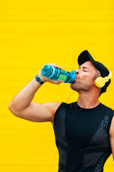 Männlicher Sportler in Sportkleidung, der Wasser aus einer Flasche trinkt und über Kopfhörer Musik hört, während er eine Trainingspause an einer gelben Wand macht - ADSF50346