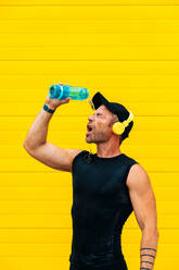 Männlicher Sportler in schwarzer Sportkleidung und Mütze mit Kopfhörern, der sich nach dem Training an einer gelben Wand Wasser aus einer Flasche ins Gesicht gießt - ADSF50347