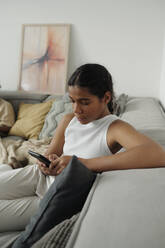 Pre-adolescent girl using smart phone sitting on sofa at home - DSHF01421