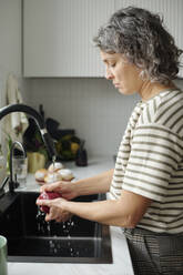 Mature woman washing apple in kitchen sink at home - DSHF01436