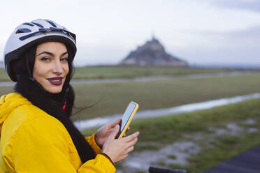 Smiling woman wearing cycling helmet with smart phone - JCCMF10967