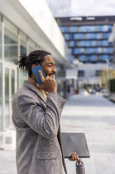 Happy businessman holding file folder and talking on smart phone at office park - IKF01487