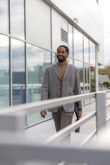 Smiling businessman wearing blazer walking on footpath near railing at office park - IKF01489