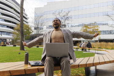 Tired businessman stretching and sitting on bench with laptop at office park - IKF01491