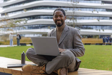Happy businessman sitting with laptop in front of office building - IKF01498