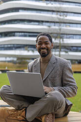 Cheerful businessman sitting with laptop on bench at office park - IKF01499