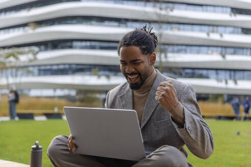 Happy businessman cheering and using laptop at office park - IKF01500