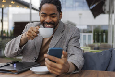 Happy businessman drinking coffee and using smart phone - IKF01507