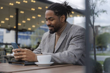 Smiling businessman sitting with coffee cup and using smart phone at sidewalk cafe - IKF01508